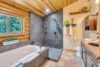 A rustic bathroom with a log cabin theme features a large bathtub next to a window with a nature view, a walk-in tiled shower, and a wooden vanity with a granite countertop and sink. The ceiling is wooden, and the floor is tiled with brown and tan squares.