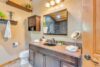 A bathroom featuring a granite countertop with a sink, a large mirror, and three overhead lights. A cabinet and shelves are on the wall, holding towels and decor items. The room also includes a toilet and a shower visible through the mirror's reflection.