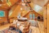 A cozy cabin bedroom with wooden walls and ceilings, featuring a bed, a small desk, two plush chairs, and colorful rugs on the wooden floor. Ceiling fans and a potted plant add to the rustic charm. French doors in the back open to a balcony with a view of nature.