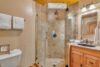 A modern bathroom with a glass-enclosed corner shower featuring textured brown tile. There's a vanity with a wooden cabinet, a rectangular mirror, and a black sink countertop. Towels are neatly hung on a nearby rack above a toilet. The walls and ceiling are wooden.
