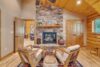 A cozy living room with wooden walls and ceiling, centered around a stone fireplace. Two padded wooden chairs face the fireplace atop a vibrant rug. To the left, a doorway reveals another room. Decorative vases and plants adorn the space for a rustic feel.