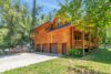 A rustic log cabin with a covered porch and three-car garage, nestled among lush trees. The home is surrounded by greenery, rocks, and a well-maintained lawn, with a clear blue sky overhead. The cabin exudes a cozy, natural charm in a serene setting.