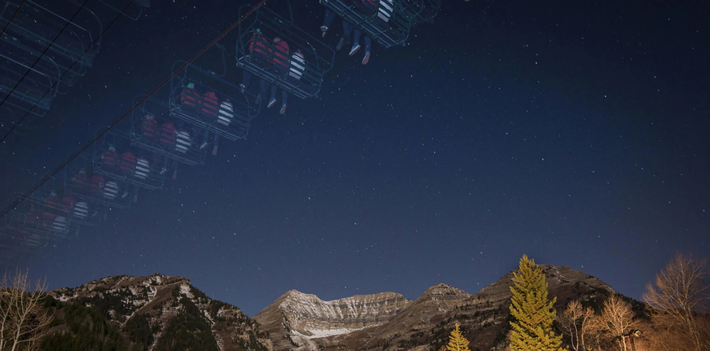 A starry night sky above a mountain range with snow-capped peaks, partially illuminated with a warm glow. Ski lift chairs are visible in the foreground, creating a sense of motion across the scene. Pine trees at the base of the mountains add to the serene atmosphere.