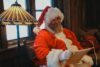 Santa Claus in a red and white suit sits in a cozy room near a window, reading a parchment scroll. A warm glow from a Tiffany-style lamp lights the scene, adding to the festive atmosphere.