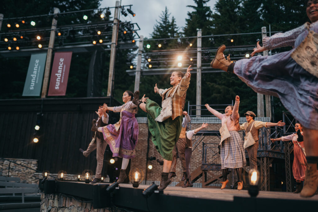 Performers in colorful period costumes dance energetically on an outdoor stage, surrounded by trees. Banners with "sundance" hang in the background, and stage lights illuminate the scene.