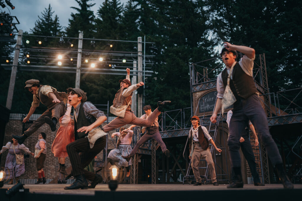 Performers in period costumes enthusiastically dance on an outdoor stage, surrounded by trees. A backdrop with scaffolding and a sign adds to the theatrical setting. Lighting illuminates the lively scene.