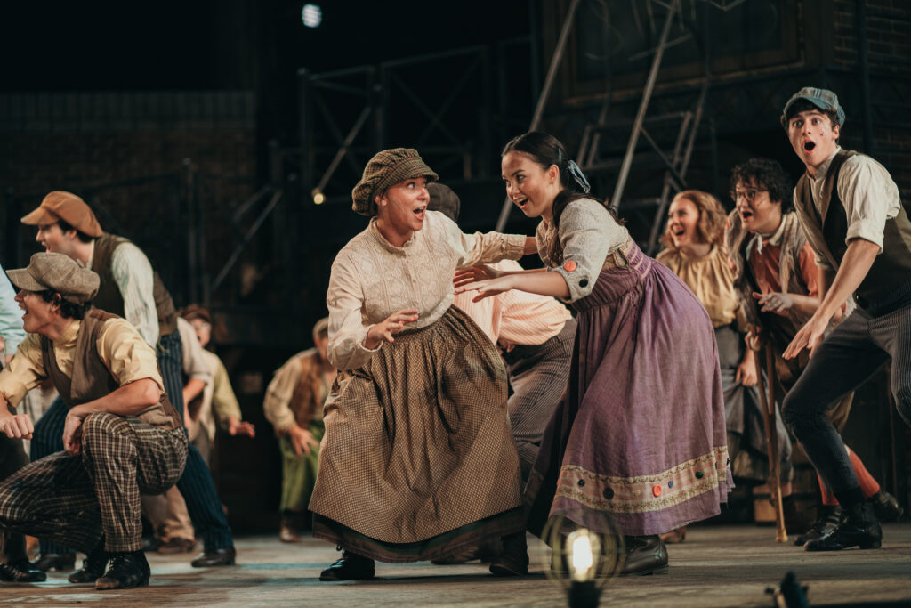 A lively stage performance features actors in period costumes. A woman in a white blouse and brown skirt gestures energetically, while another woman in a purple dress and apron smiles beside her. Other actors in caps react animatedly in the background.