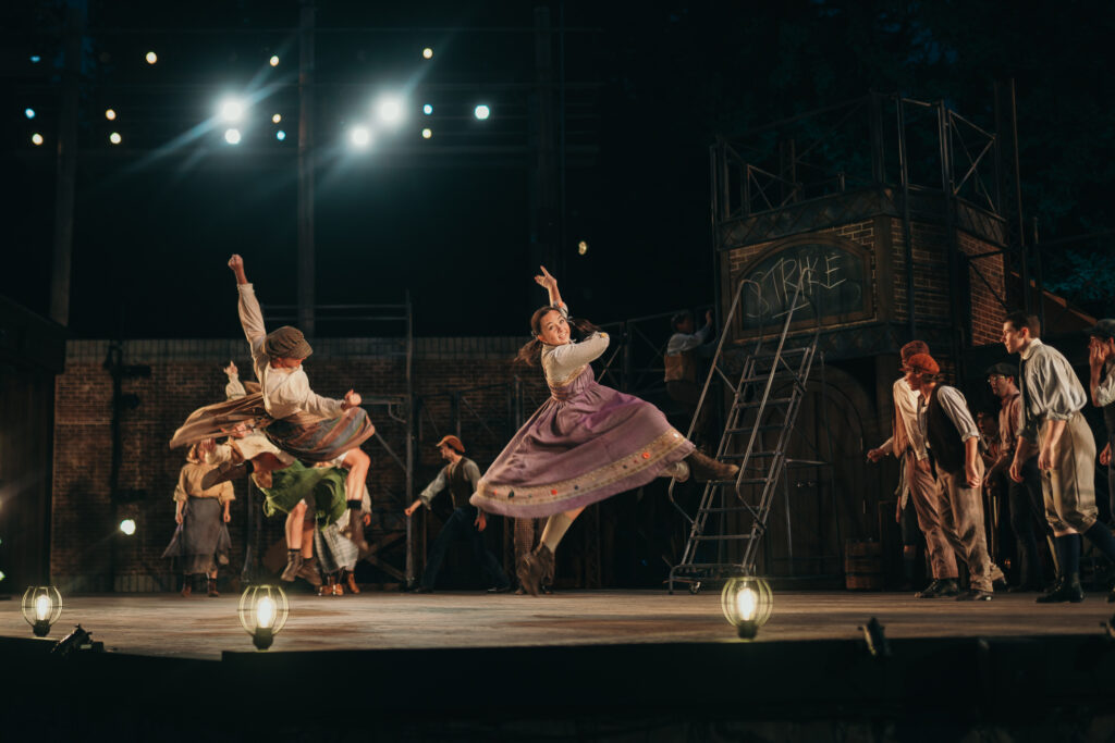 A group of performers is on an outdoor stage under bright lights. Two people in colorful costumes are leaping energetically at the front, while others in period clothing watch in the background. A ladder and some wooden structures are visible.