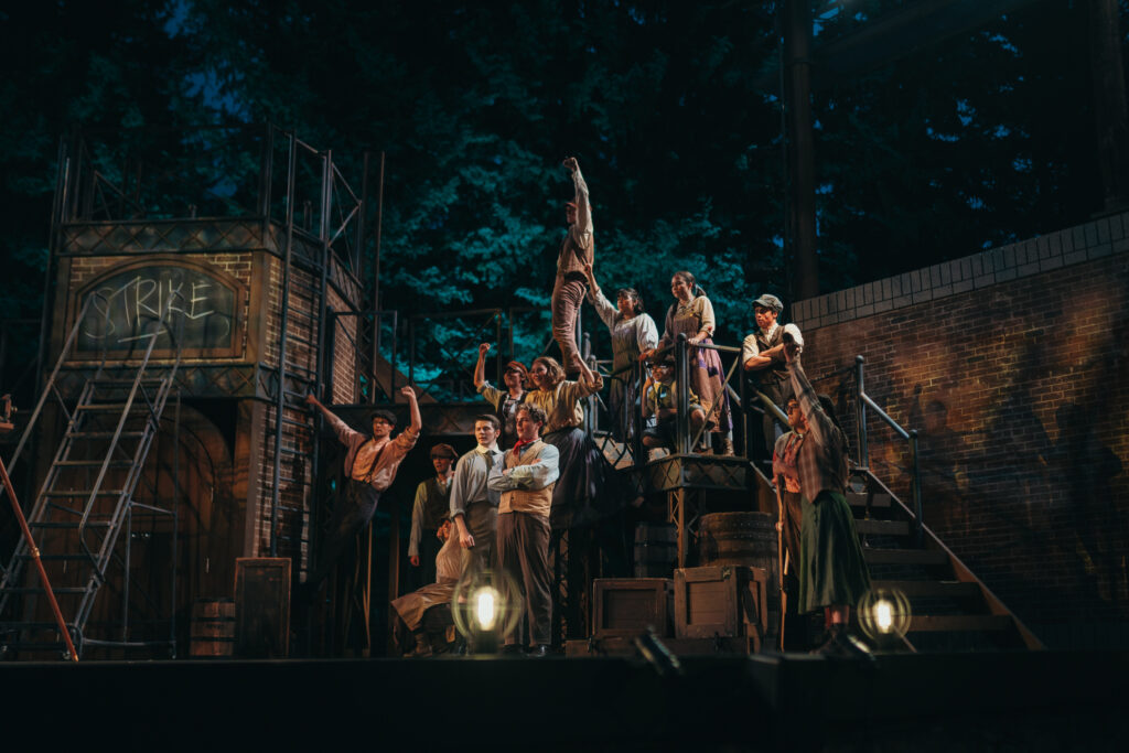 A group of actors performs on a dimly lit outdoor stage with industrial-themed scenery. One actor stands on a platform holding a newspaper, while others gather around, some pointing upwards. The backdrop features ladders and a graffitied wall.