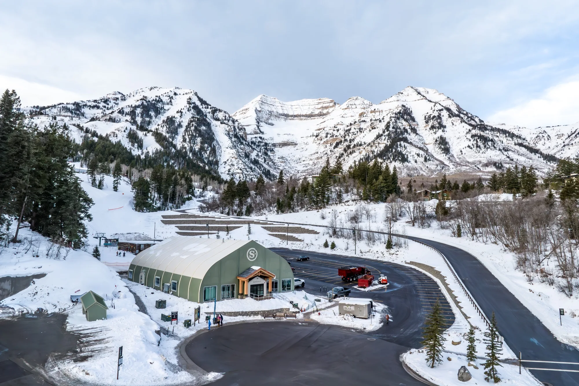 A winter scene of a ski resort with a green-roofed building surrounded by snow-covered mountains and trees. A road curves through the area, and a small crowd is visible near the entrance.