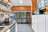 A modern kitchen with wooden walls features stainless steel appliances, open shelves, and a large window. A dining area with a table and chairs overlooks a tree-lined view. White cabinets hold a toaster and a knife set.