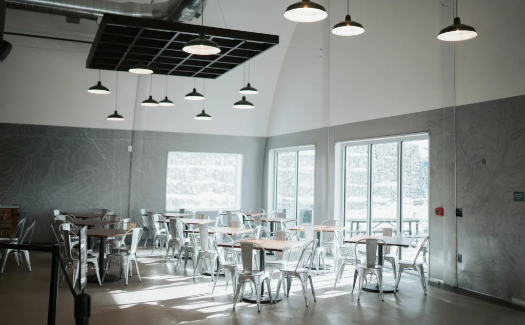 A bright, airy cafe interior with wooden tables and metal chairs. Large windows allow natural light to flood the space. Several pendant lights hang from the high ceiling. The atmosphere is modern and inviting.
