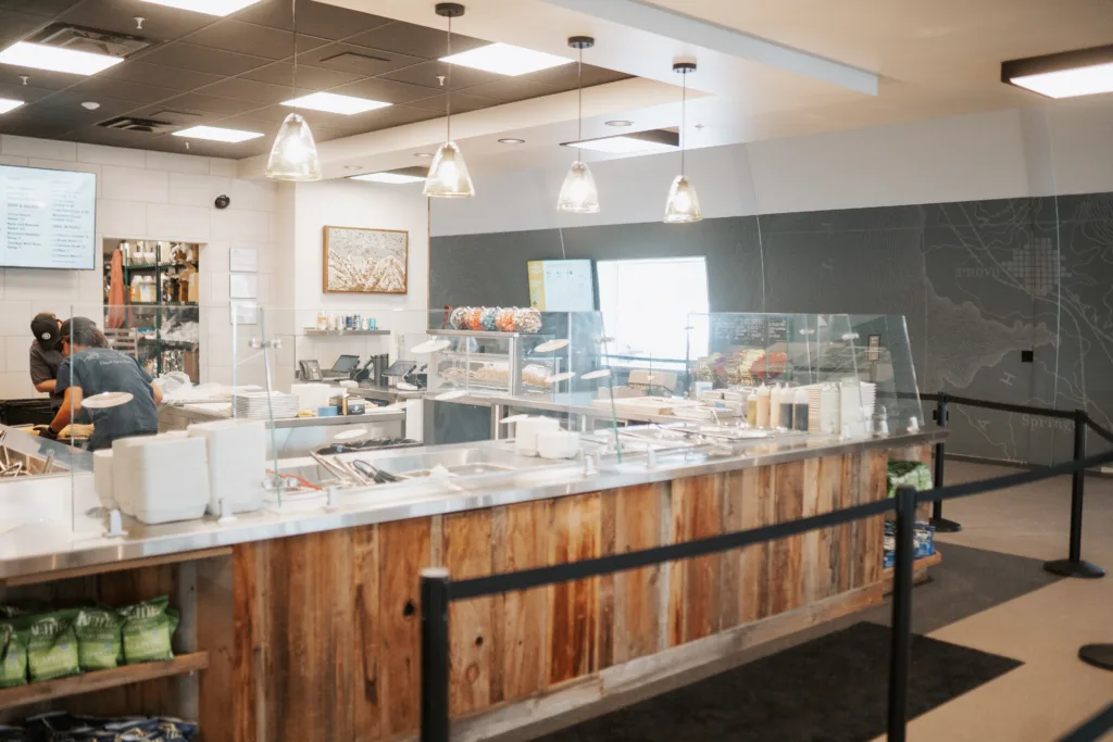 A modern deli with a wooden counter and glass display. Various food items like sandwiches and salads are visible. Hanging pendant lights illuminate the area. A person is working behind the counter. Menu boards are on the wall.