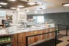 A modern deli with a wooden counter and glass display. Various food items like sandwiches and salads are visible. Hanging pendant lights illuminate the area. A person is working behind the counter. Menu boards are on the wall.