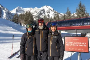 Three people in winter gear stand smiling on a snowy mountain slope. Behind them is a ski lift, snowy trees, and mountain peaks. A sign reads, 