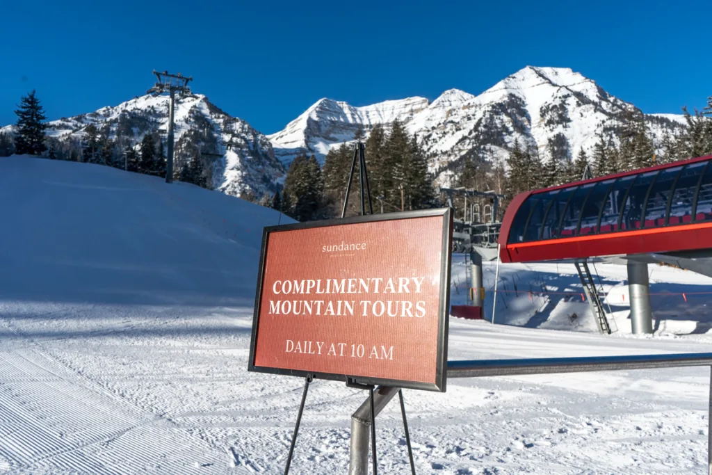A sign reads "Complimentary Mountain Tours Daily at 10 AM" in front of a ski lift and snow-covered mountains under a clear blue sky. Snowy trees and a red ski lift are visible in the background.