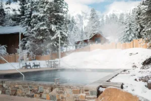 A steaming outdoor pool is set in a snowy landscape with snow-covered trees and mountains, perfect for those exploring Utah vacation packages. Steam rises from the water, enclosed by a wooden fence. Nearby, a small rustic cabin invites you to unwind after enjoying Sundance dining adventures.