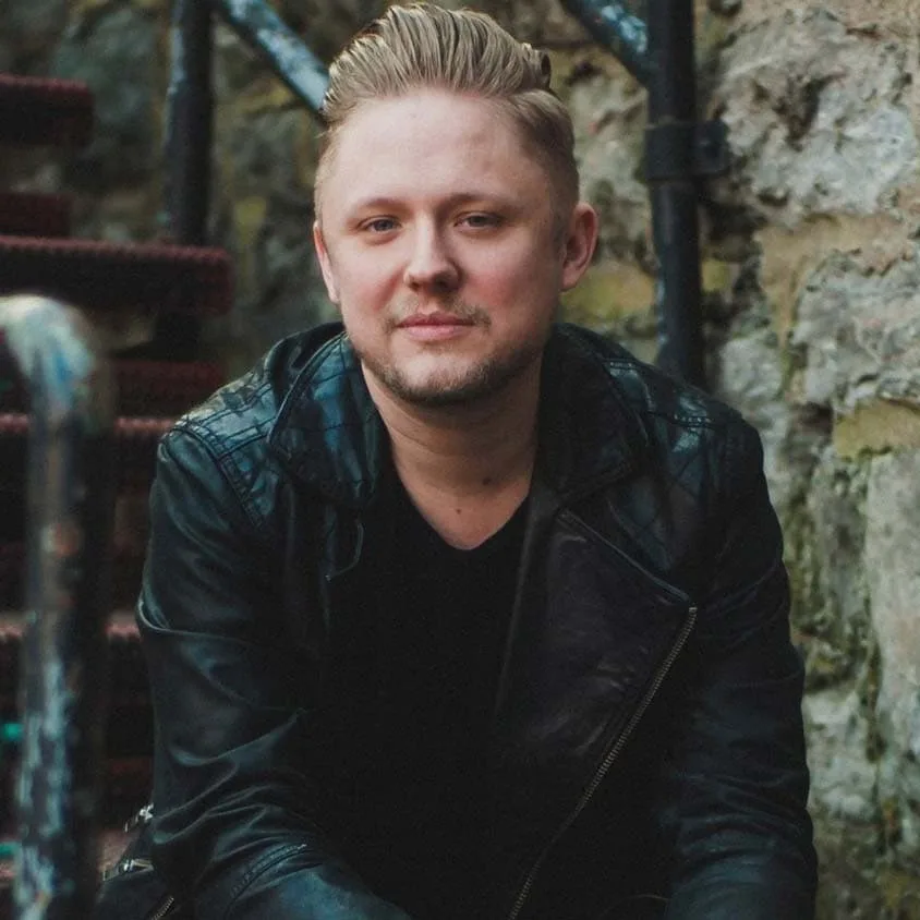 A person with short blond hair and a trimmed beard, wearing a black leather jacket and black shirt, sits outdoors on a rustic staircase reminiscent of the charm of the Bluebird Cafe. The background consists of stone walls and a metal railing.