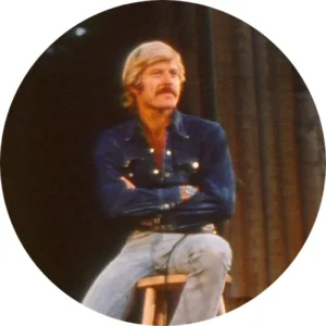 A person with blonde hair and a mustache sits on a stool, arms crossed, wearing a denim shirt and jeans. The background is dark and slightly blurred.