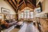 A rustic living room with a high wooden ceiling and exposed beams. It features a leather sofa, wooden coffee table, and stone fireplace. Large windows provide natural light, and a round dining table with chairs sits by the window. A painting is on the wall.