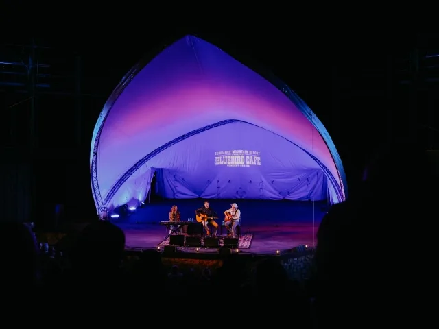 Two musicians perform on stage under a large, illuminated canopy at night. The words "Bluebird Cafe" glow on the backdrop, evoking the charm of Sundance dining. The stage is set with a guitar, keyboard, and microphones while the audience watches in silhouette.