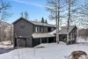 A modern gray house with large windows and a metal roof stands against a snowy landscape. As the moonrise casts a gentle glow, bare trees encircle the home. A driveway winds to an attached garage, crowned by a circular window. The sky is clear with hints of blue.