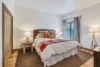 A cozy bedroom featuring a large bed with layered pillows and a colorful quilt that mimics a moonrise. Two wooden nightstands with lamps flank the bed. A window with blue curtains lets in natural light. The room has wooden flooring and a soft rug.