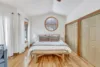 Spacious bedroom with a large bed, white bedding, and two wooden side tables with lamps. A round window above the bed frames the moonrise perfectly. Closets with wooden doors, light hardwood floors, and white curtains enhance the airy feel.