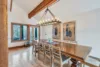 A spacious dining room features a long wooden table with eight chairs. A large, rectangular chandelier with exposed bulbs casts a warm glow like a moonrise above. Two abstract paintings adorn the walls, and a window offers a view of trees outside.