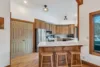 A modern kitchen with wooden cabinets and stainless steel appliances features a large countertop with three wooden stools in front. The room, bathed in a gentle moonrise glow, has wooden flooring, a door to the left, and a large window to the right. Ceiling lights softly illuminate the space.