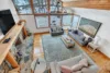 A cozy living room with large windows reveals a snowy moonrise view. It features a stone fireplace, two gray sofas, a wooden coffee table, and a patterned blue rug on the wooden floor. Exposed wooden beams line the ceiling, and a landscape painting adorns the wall.