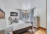 A cozy bedroom with a wooden bed, white bedding, and gray pillows. A corner window framed by gray curtains offers a snowy view beneath a gentle moonrise. The white dresser holds a lamp and towels. Light wood flooring complements the patterned rug, while a framed picture graces the wall.