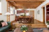 A cozy living room with wooden beams on the ceiling, a green couch, and a wooden coffee table with plants. The dining area has a table with chairs and a chandelier. A kitchen with red cabinets and modern appliances is in the background.