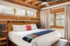 A cozy bedroom with a rustic log cabin design features a large bed with white linens, accented by a colorful striped throw. Two windows bring in natural light, and a ceiling fan and wooden beams add to the warm, inviting atmosphere.