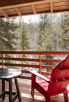 A cozy wooden balcony with a bright red Adirondack chair and a small round table overlooks a peaceful winter forest scene. Snow blankets the ground, and tall evergreen trees stand under a partly cloudy sky.