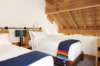 Cozy attic bedroom with two single beds, each with white linens and a blue blanket. One blanket features a colorful stripe design. A black lamp sits between the beds, and the ceiling has exposed wooden beams.