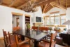 A rustic dining room with a dark wood table and six leather chairs. Wall-mounted antler chandelier hangs from a wooden beamed ceiling. A fireplace and TV adorn the back wall. Large windows let in natural light. Cozy seating area visible in the background.