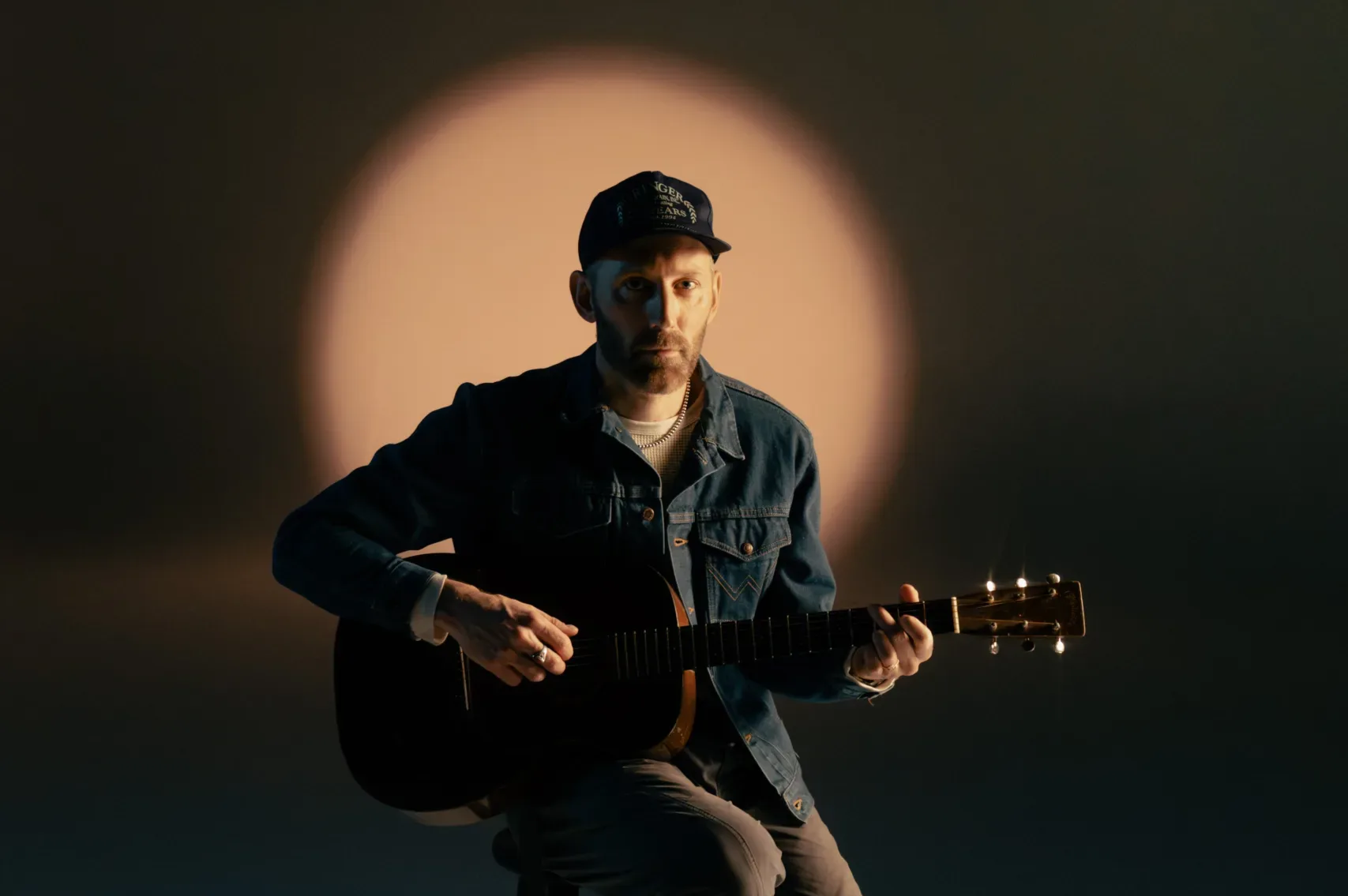 Mat Kearney sits on a stool playing an acoustic guitar. He wears a dark jacket, cap, and light-colored sneakers. The background is dim, with a circular light creating a halo effect behind him on a neutral backdrop.