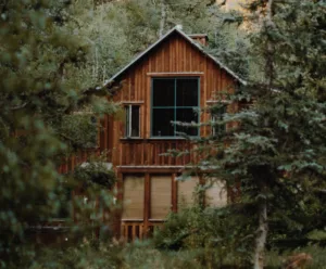 A rustic, wooden cabin partially obscured by dense trees offers the perfect retreat. With large windows showcasing lush greenery, it promises seclusion and tranquility for those exploring Utah vacation packages or indulging in nearby Sundance dining for a complete retreat experience.