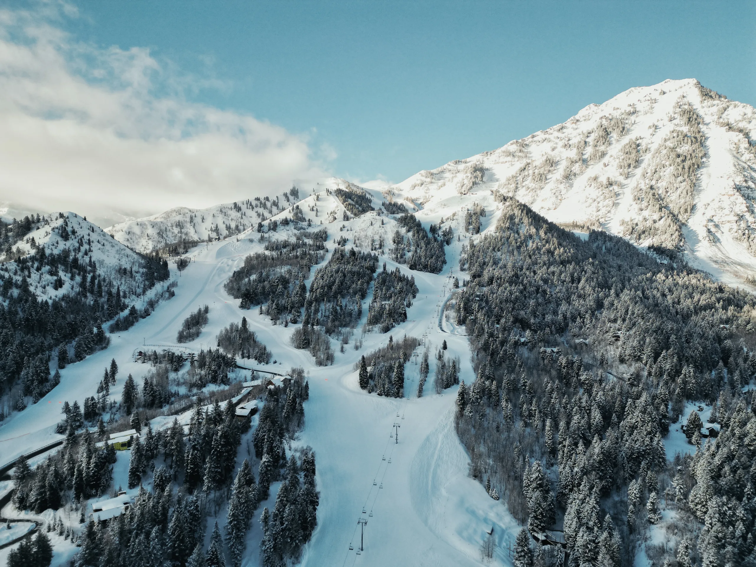 A snow-covered mountain landscape with ski slopes, surrounded by dense forests. Lift lines are visible on the slopes. The sky is clear with a few clouds, enhancing the winter scenery.