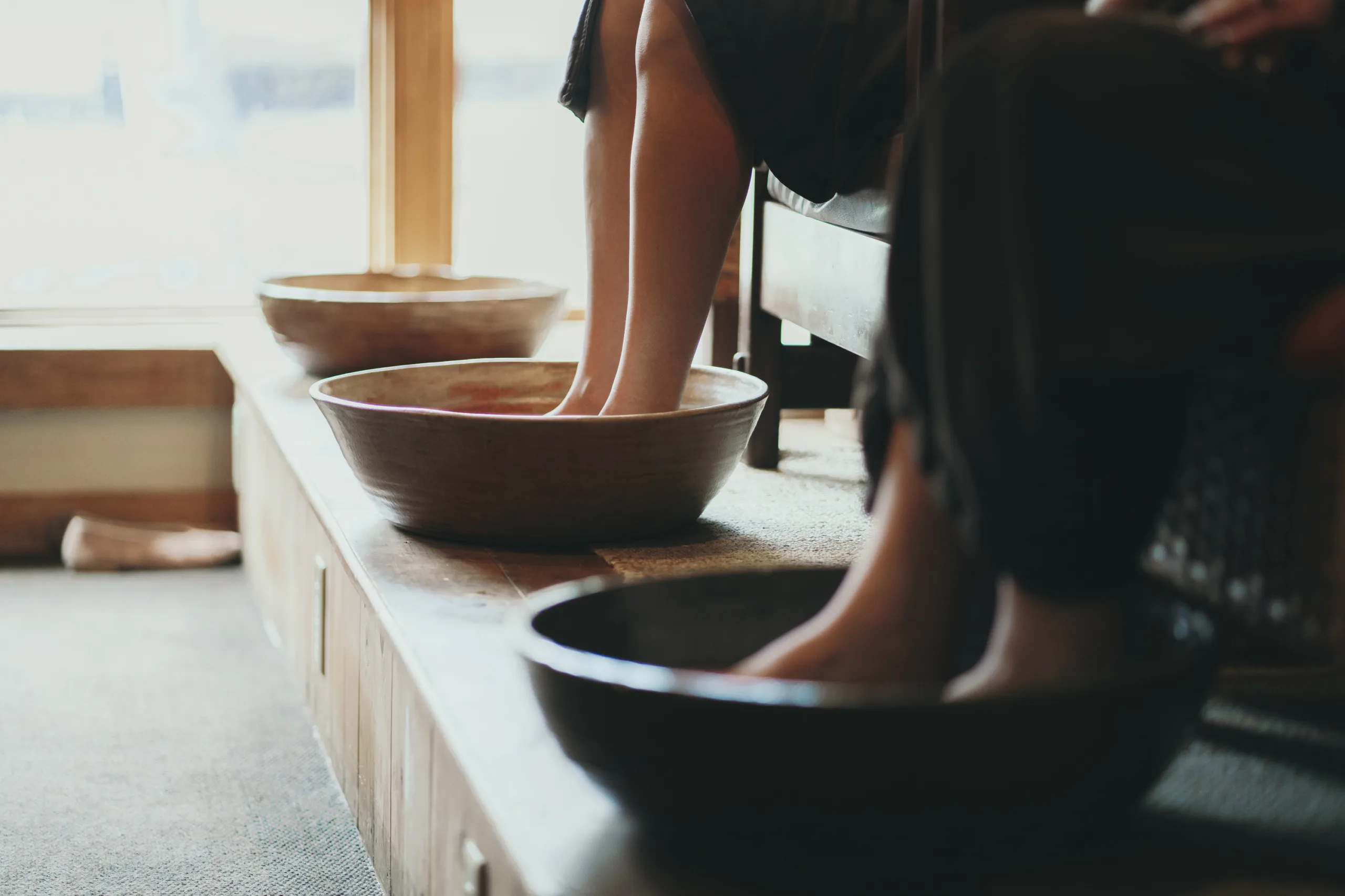 People soaking their feet in wooden bowls filled with water, sitting on a raised platform in a serene setting. Natural light streams through a window, creating a calm and relaxing ambiance.