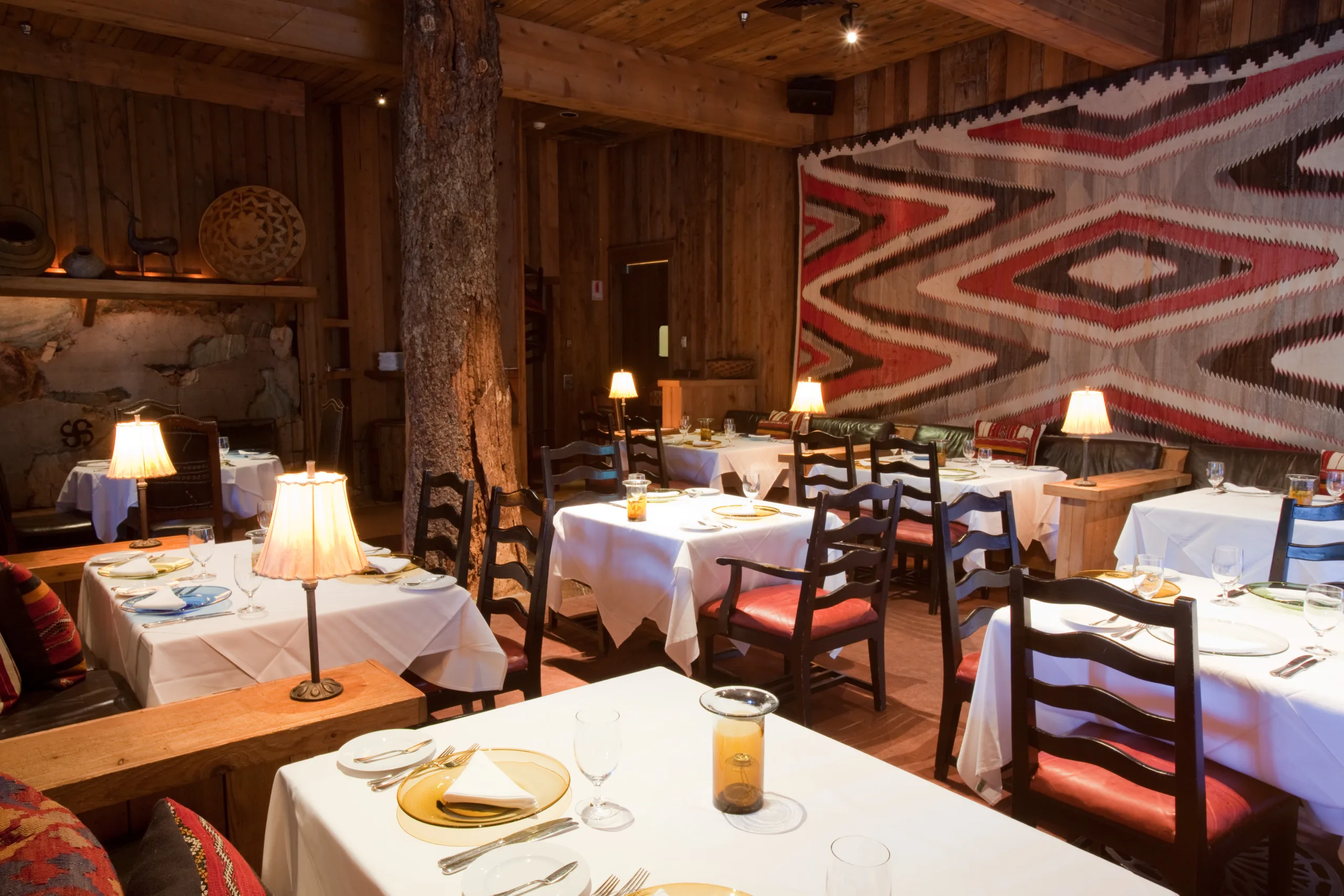 A cozy restaurant interior with wooden walls and ceiling, decorated with a large geometric patterned tapestry. White tablecloths cover the tables, set with plates and glasses. Lamps on each table create a warm, inviting atmosphere.