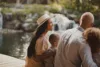 A family of four sits by a tranquil pond with a gentle waterfall in the background, enjoying their Utah vacation. The woman wears a white hat and light dress, while the man and children face the water. Lush greenery surrounds them, creating a peaceful atmosphere reminiscent of Sundance dining experiences.
