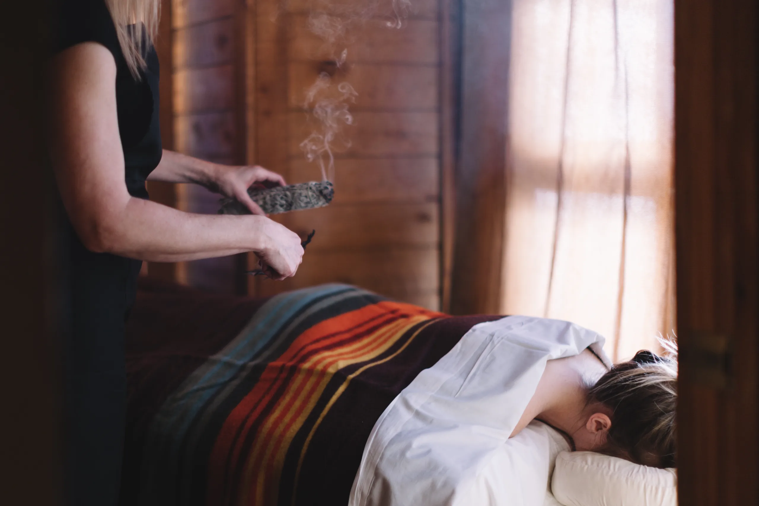 A person lies face down on a massage table, covered with a striped blanket. Another person holds a smoking bundle of sage near the table, in a cozy, wooden room with soft light coming through curtains.