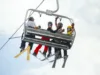A group of five people sitting on a ski lift, equipped with skis and snowboards. They are dressed in colorful winter apparel, chatting and enjoying the ride against a cloudy sky backdrop.