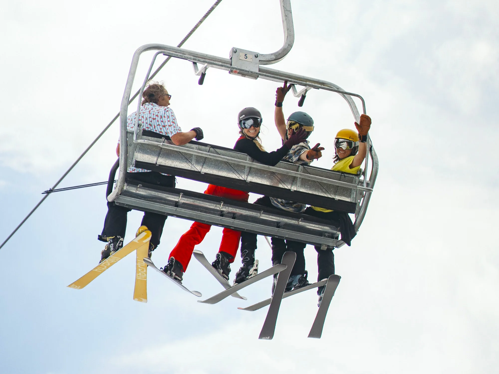 A group of five people sitting on a ski lift, equipped with skis and snowboards. They are dressed in colorful winter apparel, chatting and enjoying the ride against a cloudy sky backdrop.