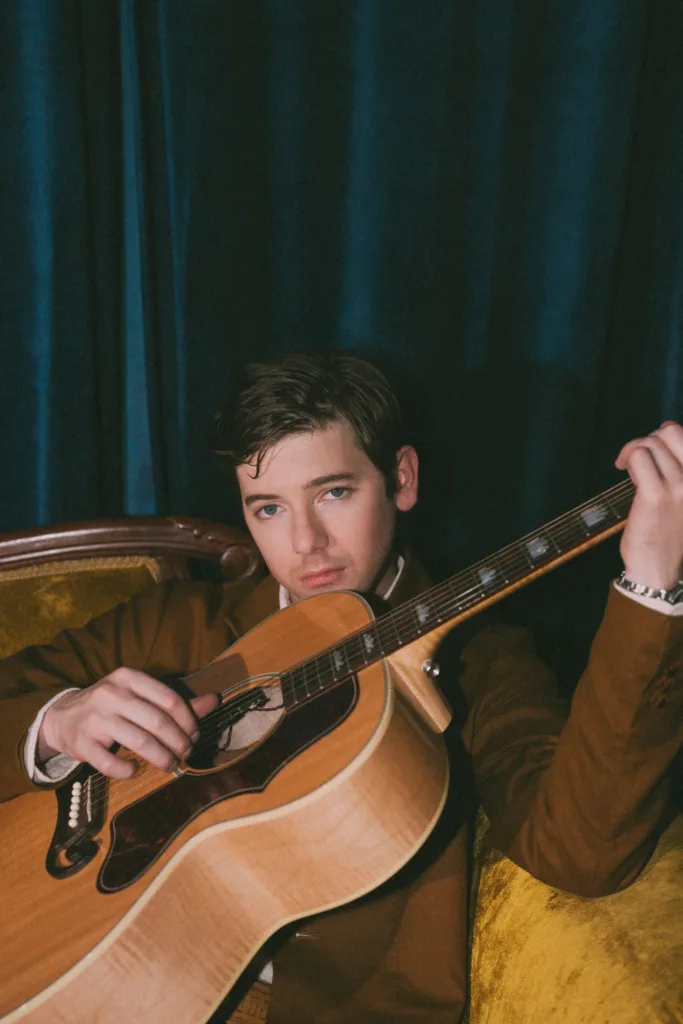 A person sits thoughtfully on a vintage chair at the Bluebird Cafe, gently resting one hand on the acoustic guitar strings. The scene is framed by a dark blue curtain, and warm light bathes everything in a comforting glow.