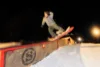 A snowboarder performs a trick on a rail in a snowy terrain park at night, illuminated by bright lights. The sky is dark, and the snow glows under the artificial lighting, highlighting the motion and energy of the scene.