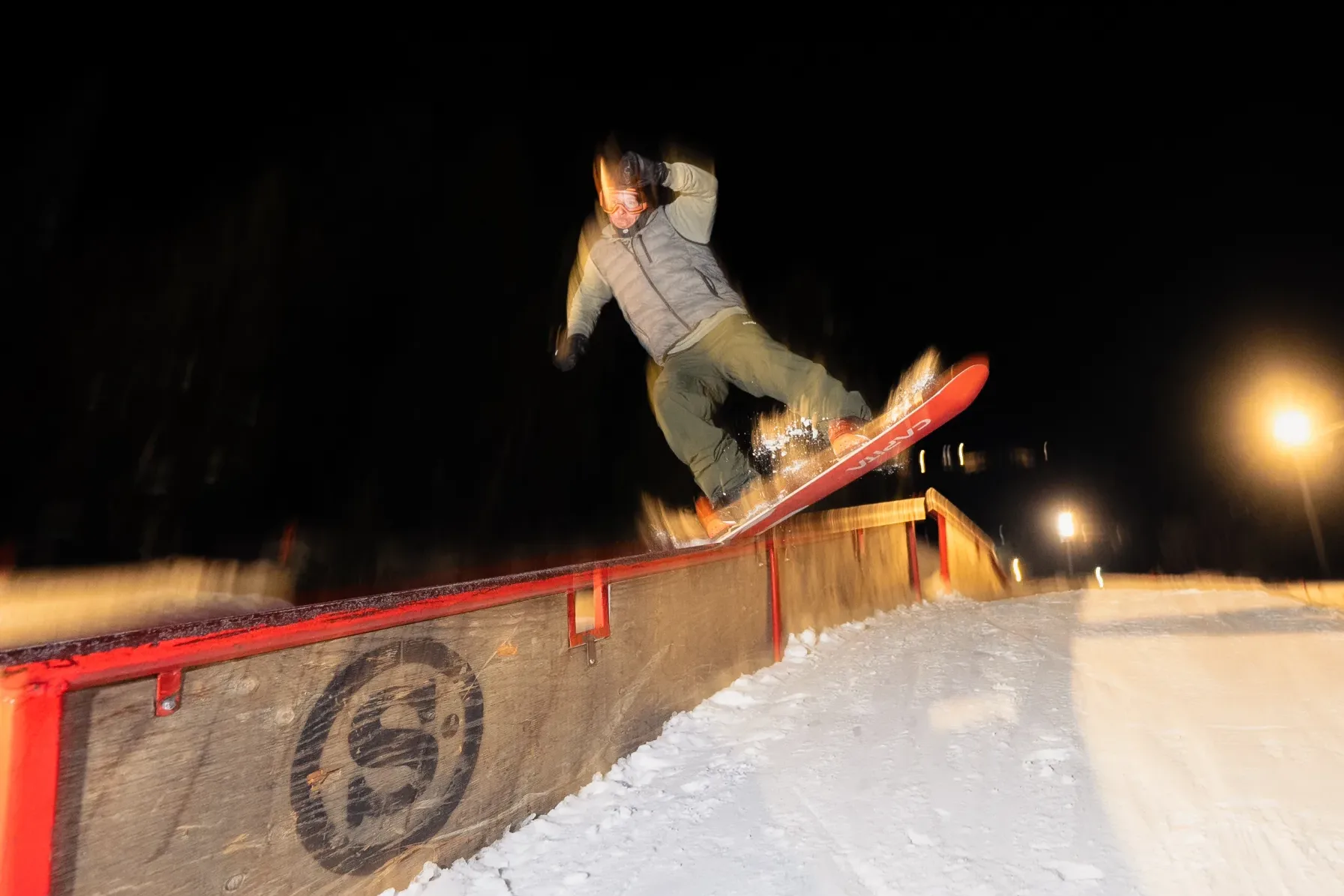 A snowboarder performs a trick on a rail in a snowy terrain park at night, illuminated by bright lights. The sky is dark, and the snow glows under the artificial lighting, highlighting the motion and energy of the scene.
