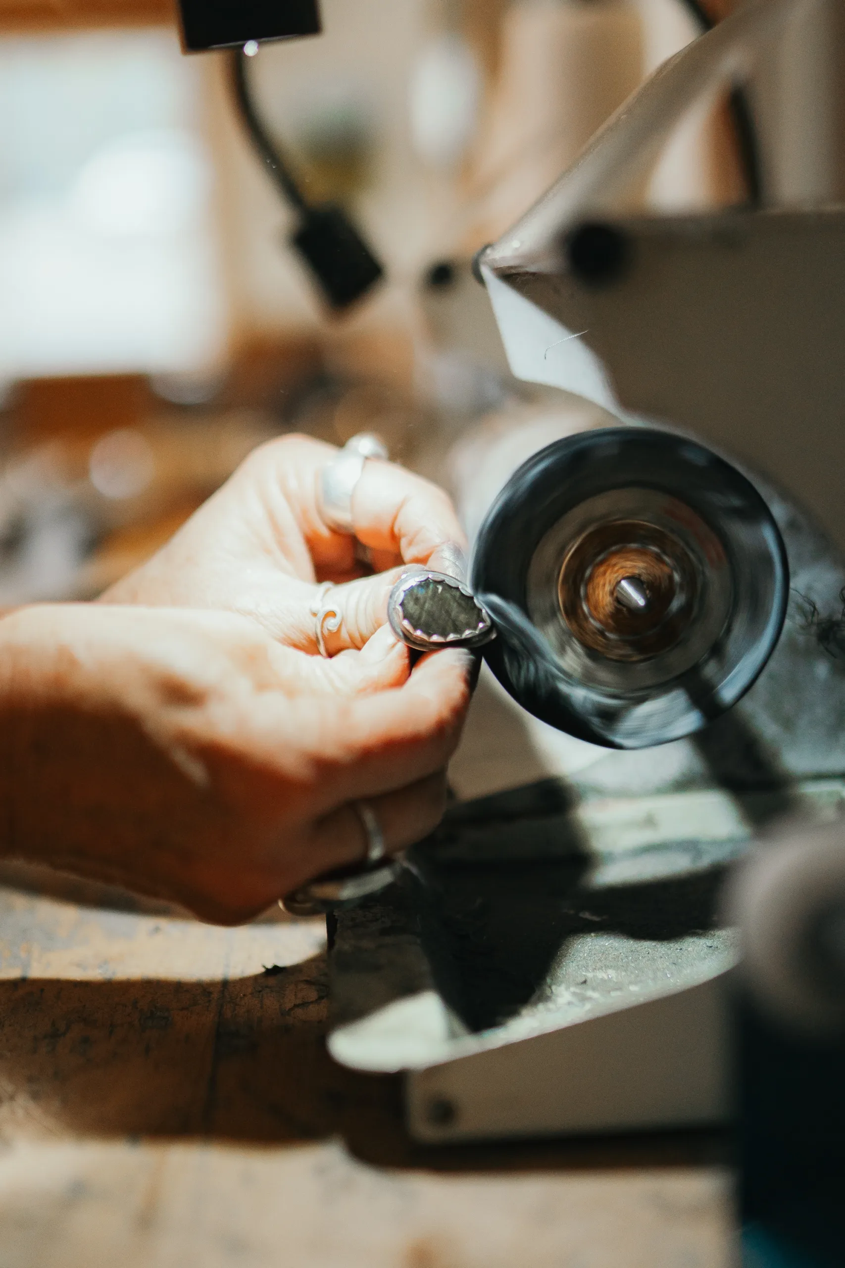 A person wearing rings is holding a ring against a spinning polishing wheel, working on its surface. The surroundings are slightly blurred, focusing on the hand and the tool. The scene suggests jewelry crafting or metalwork.